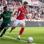 Twee voetballers in rood-witte en groene wedstrijdkleding betwisten de bal langs de zijlijn.