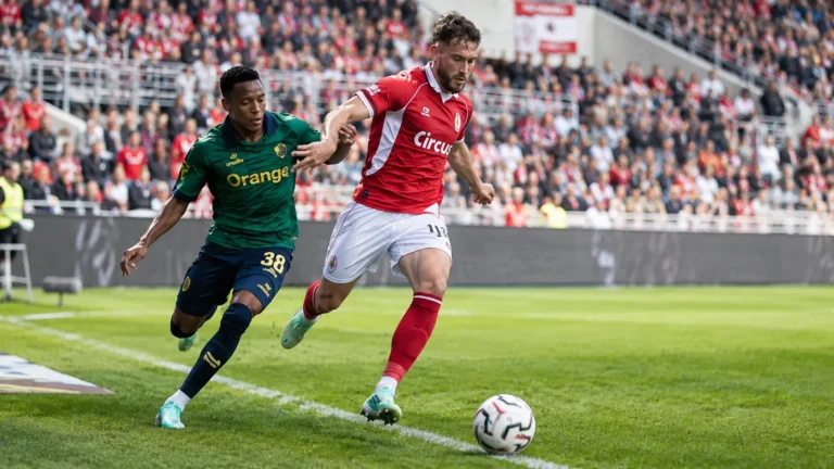 Twee voetballers in rood-witte en groene wedstrijdkleding betwisten de bal langs de zijlijn.