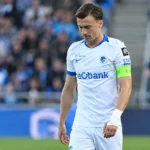Een KRC Genk-speler in een wit tenue kijkt naar beneden op het veld.