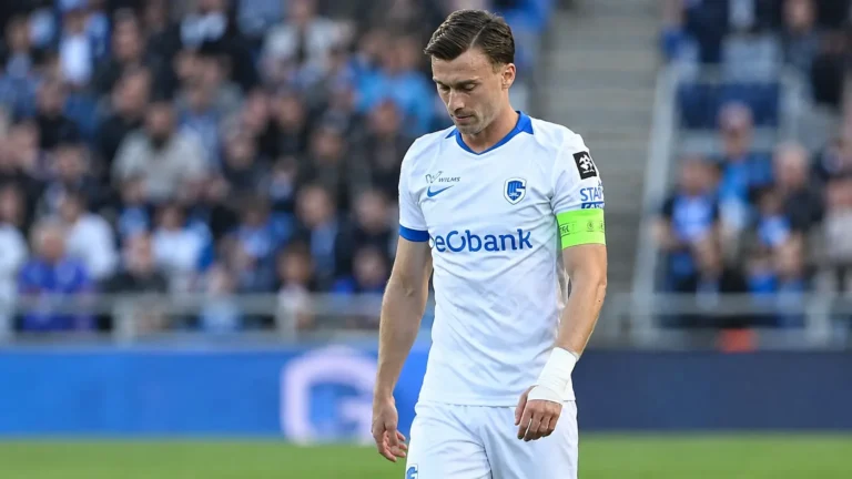 Een KRC Genk-speler in een wit tenue kijkt naar beneden op het veld.