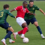 Voetballers in een rood en een donkergroen tenue in een duel om de bal.