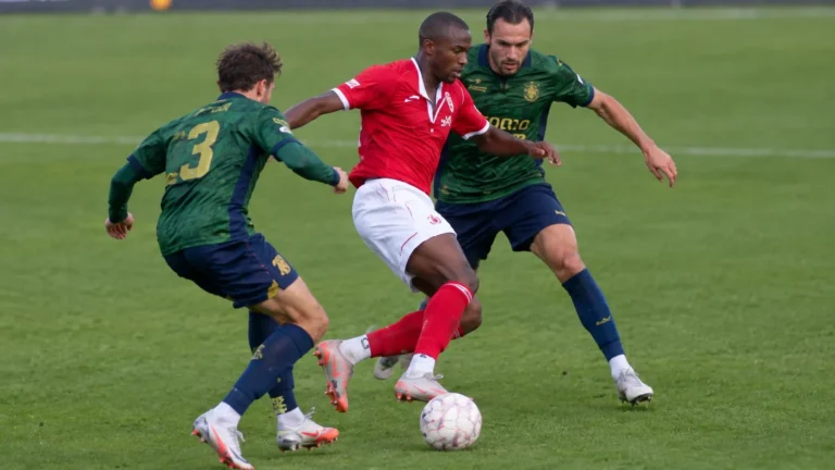 Voetballers in een rood en een donkergroen tenue in een duel om de bal.