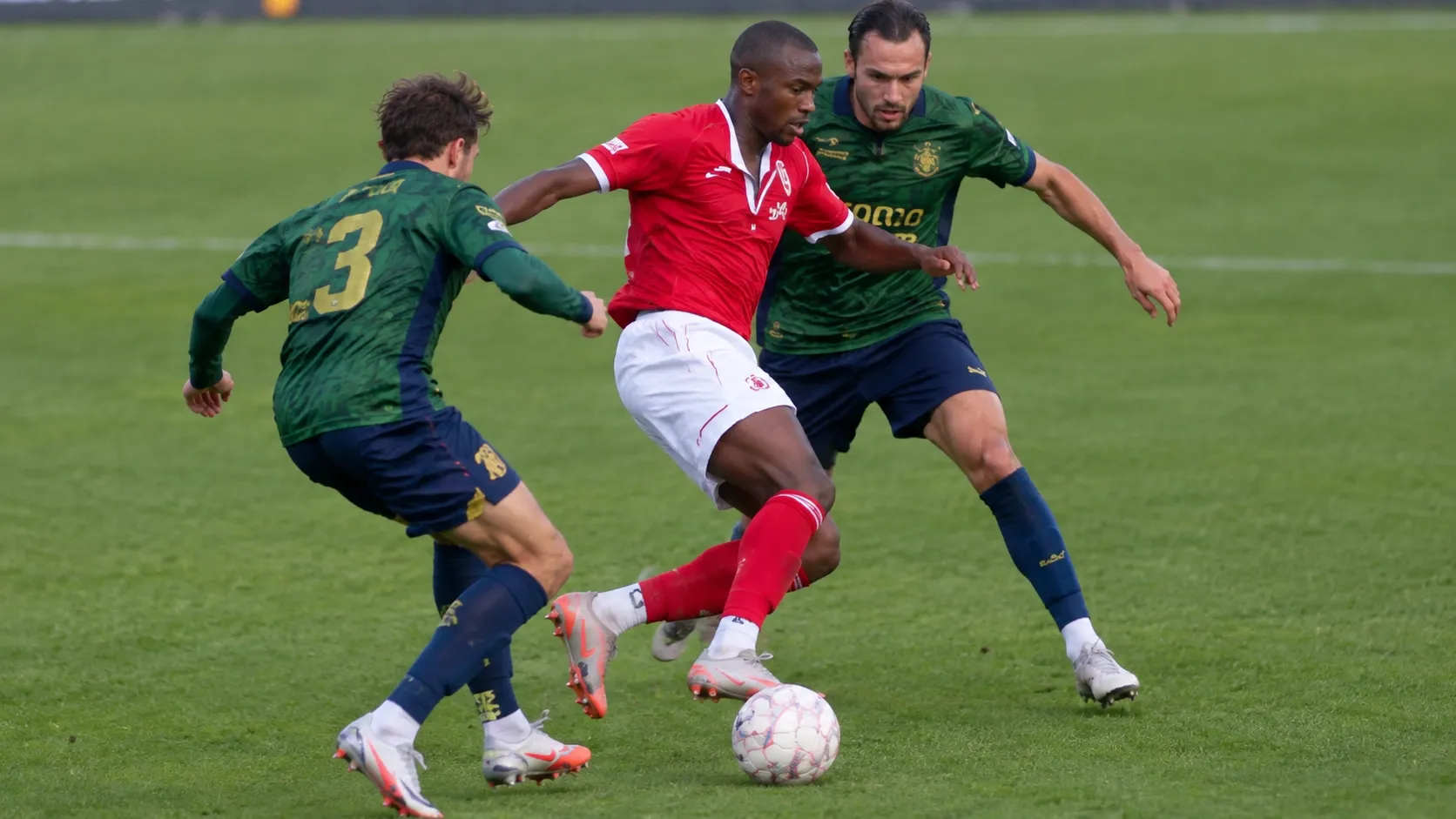 Voetballers in een rood en een donkergroen tenue in een duel om de bal.
