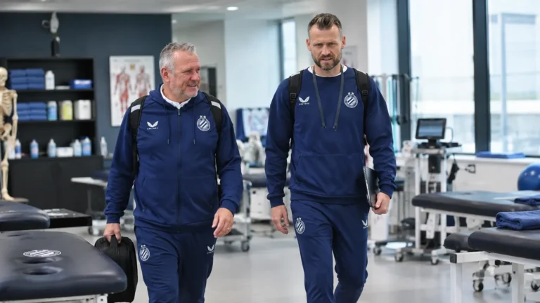 Twee medewerkers in donkere clubtrainingspakken in een medische behandelruimte.