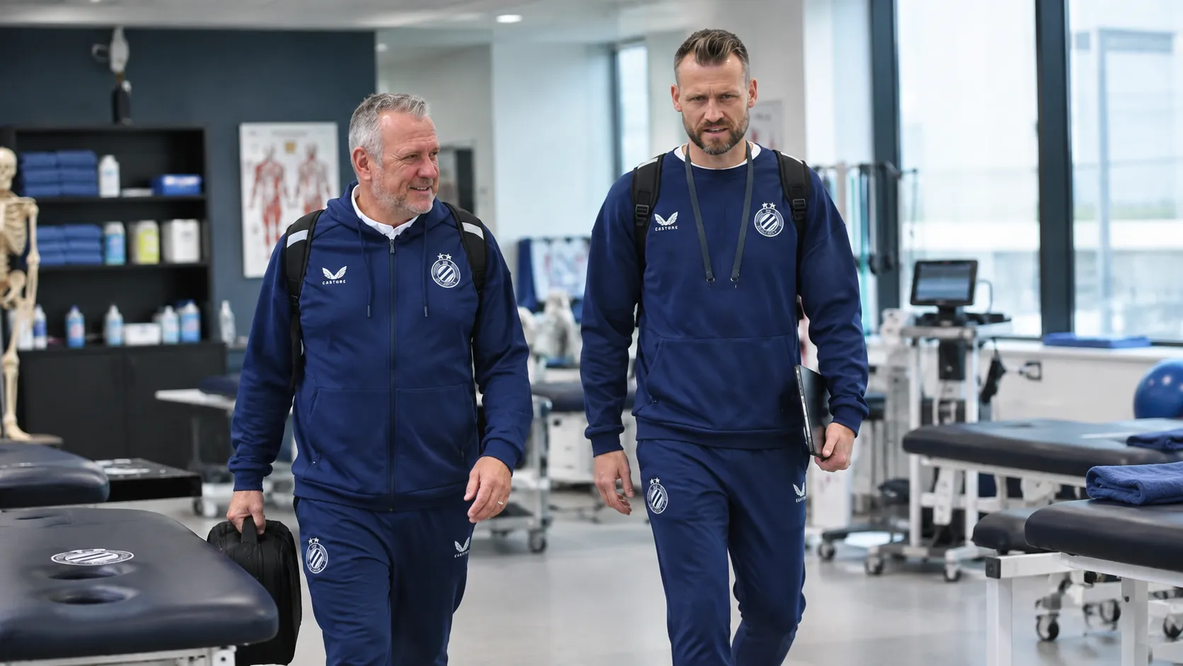 Twee medewerkers in donkere clubtrainingspakken in een medische behandelruimte.