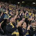 Een collectieve groep Union Saint-Gilloise-supporters applaudisseert met geel-blauwe sjaals op de tribune.