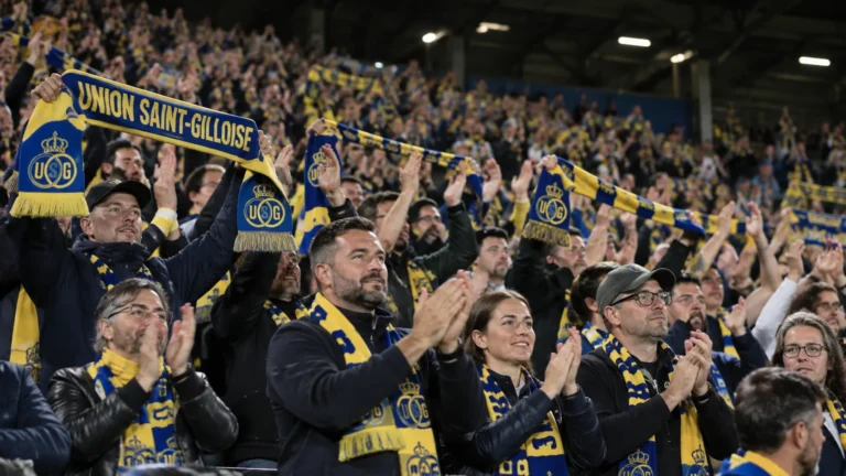 Een collectieve groep Union Saint-Gilloise-supporters applaudisseert met geel-blauwe sjaals op de tribune.