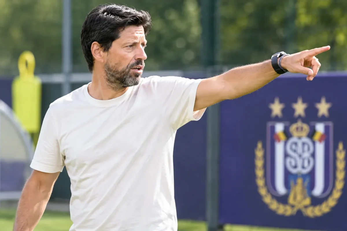 Een Anderlecht-coach geeft aanwijzingen naast een trainingsveld met een clubembleem op een vast bord.