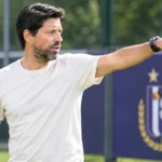 Een Anderlecht-coach geeft aanwijzingen naast een trainingsveld met een clubembleem op een vast bord.