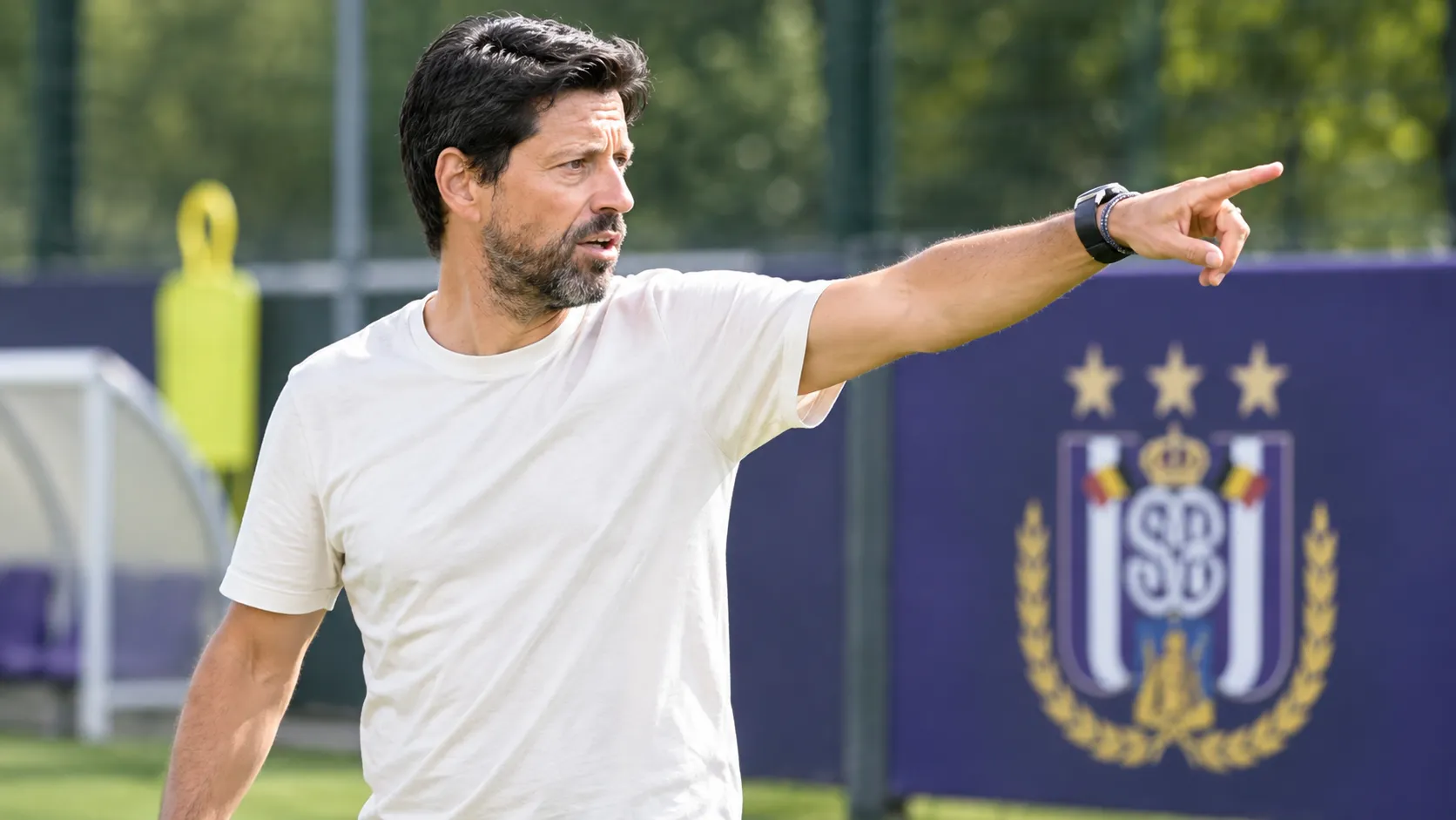 Een Anderlecht-coach geeft aanwijzingen naast een trainingsveld met een clubembleem op een vast bord.