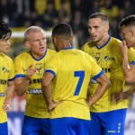 STVV-spelers in gele shirts overleggen samen op het veld tijdens een thuiswedstrijd.