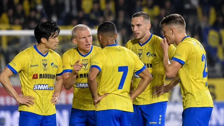 STVV-spelers in gele shirts overleggen samen op het veld tijdens een thuiswedstrijd.