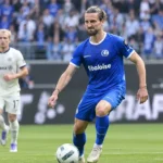 Een voetballer in een blauw KAA Gent-tenue staat op het veld, met een tweede speler op de achtergrond.