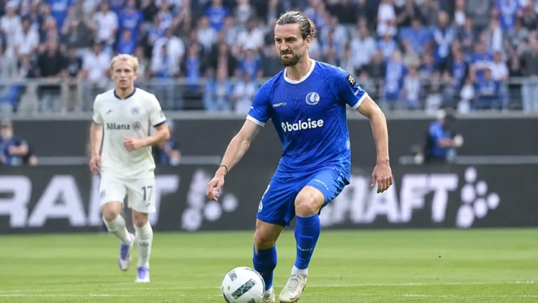 Een voetballer in een blauw KAA Gent-tenue staat op het veld, met een tweede speler op de achtergrond.