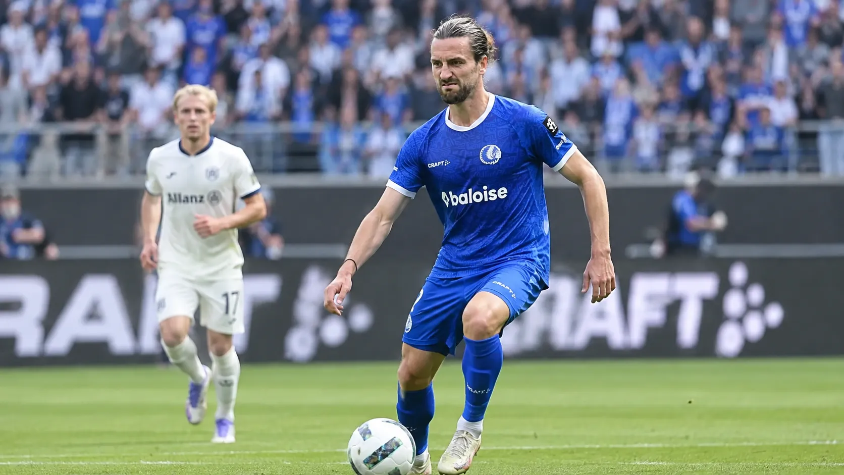 Een voetballer in een blauw KAA Gent-tenue staat op het veld, met een tweede speler op de achtergrond.