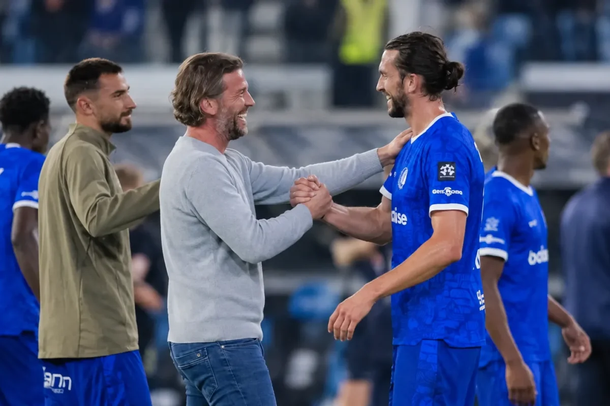 Een staflid en spelers in blauw wisselen elkaar een handdruk uit naast het veld.