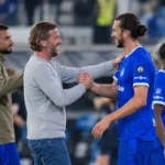 Een staflid en spelers in blauw wisselen elkaar een handdruk uit naast het veld.
