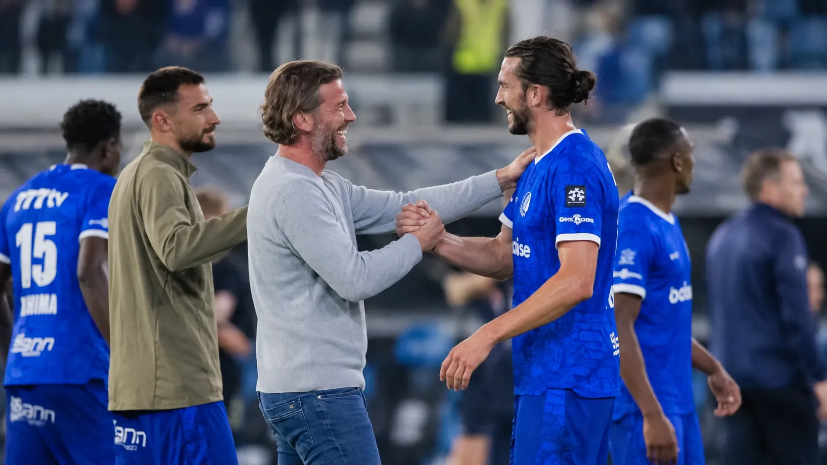 Een staflid en spelers in blauw wisselen elkaar een handdruk uit naast het veld.