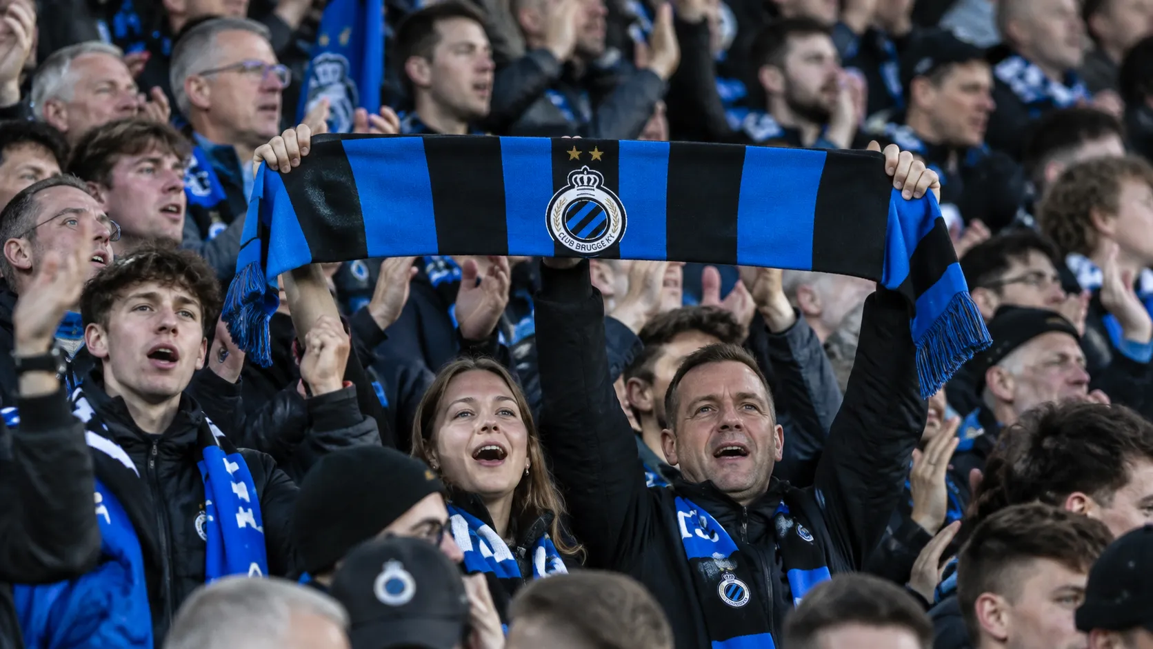 Een collectieve groep Club Brugge-supporters met één blauw-zwarte sjaal in de tribune.