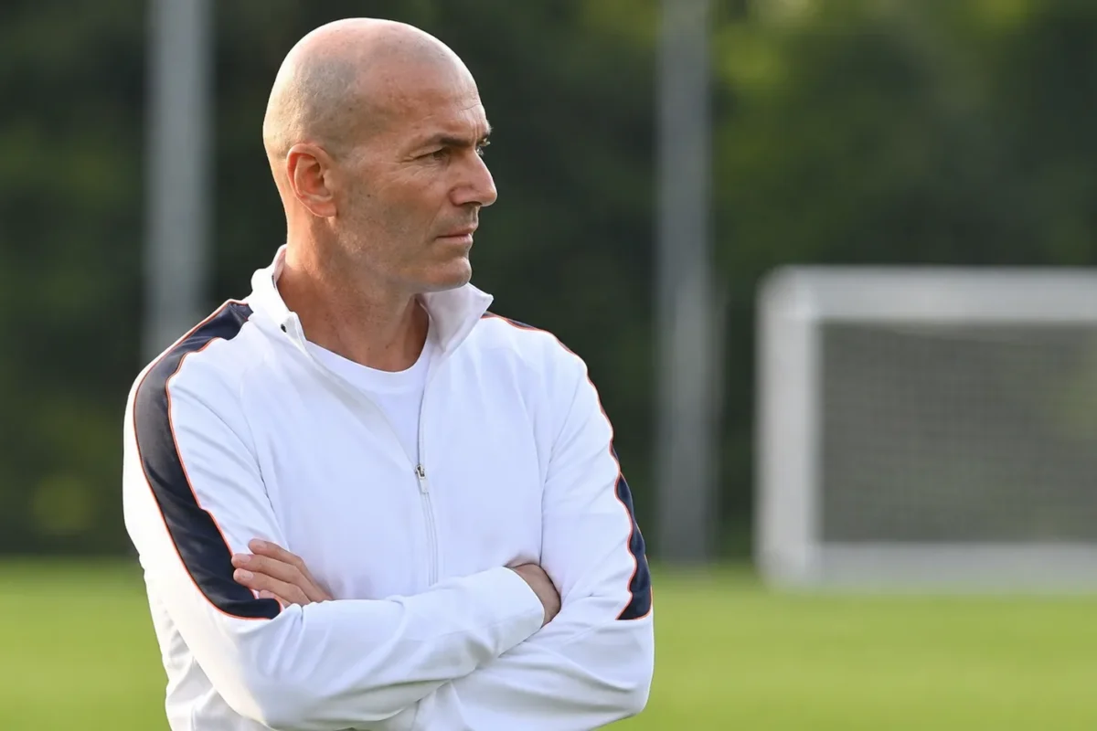 Een coach in een witte trainingsjas observeert een oefensessie langs het trainingsveld.
