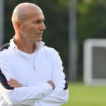 Een coach in een witte trainingsjas observeert een oefensessie langs het trainingsveld.