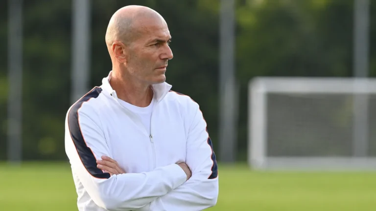 Een coach in een witte trainingsjas observeert een oefensessie langs het trainingsveld.