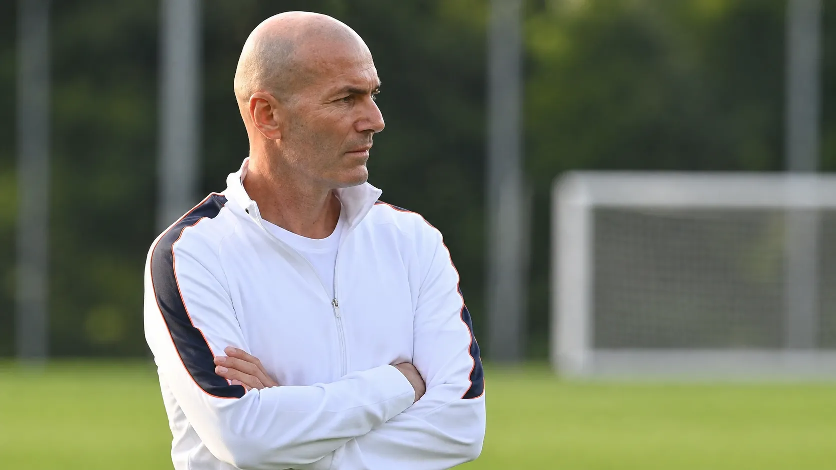 Een coach in een witte trainingsjas observeert een oefensessie langs het trainingsveld.