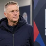 Een voetbalcoach in neutrale jas naast een Nations League-evenementbord aan een stadion.