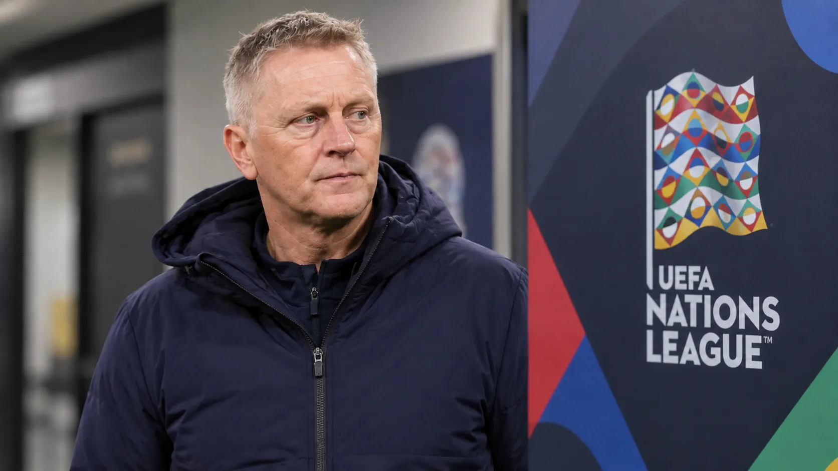 Een voetbalcoach in neutrale jas naast een Nations League-evenementbord aan een stadion.