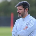 Een bondscoach in een licht trainingsjack observeert aandachtig een trainingsveld.