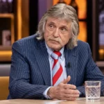 Johan Derksen aan een talkshowtafel in een televisiestudio.