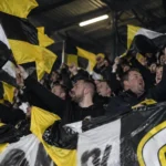 Supporters zwaaien met zwart-gele vlaggen in een voetbaltribune.