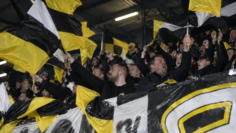 Supporters zwaaien met zwart-gele vlaggen in een voetbaltribune.