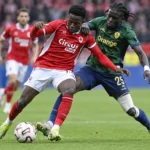 Twee voetballers in rode en groene wedstrijdkledij strijden om de bal.