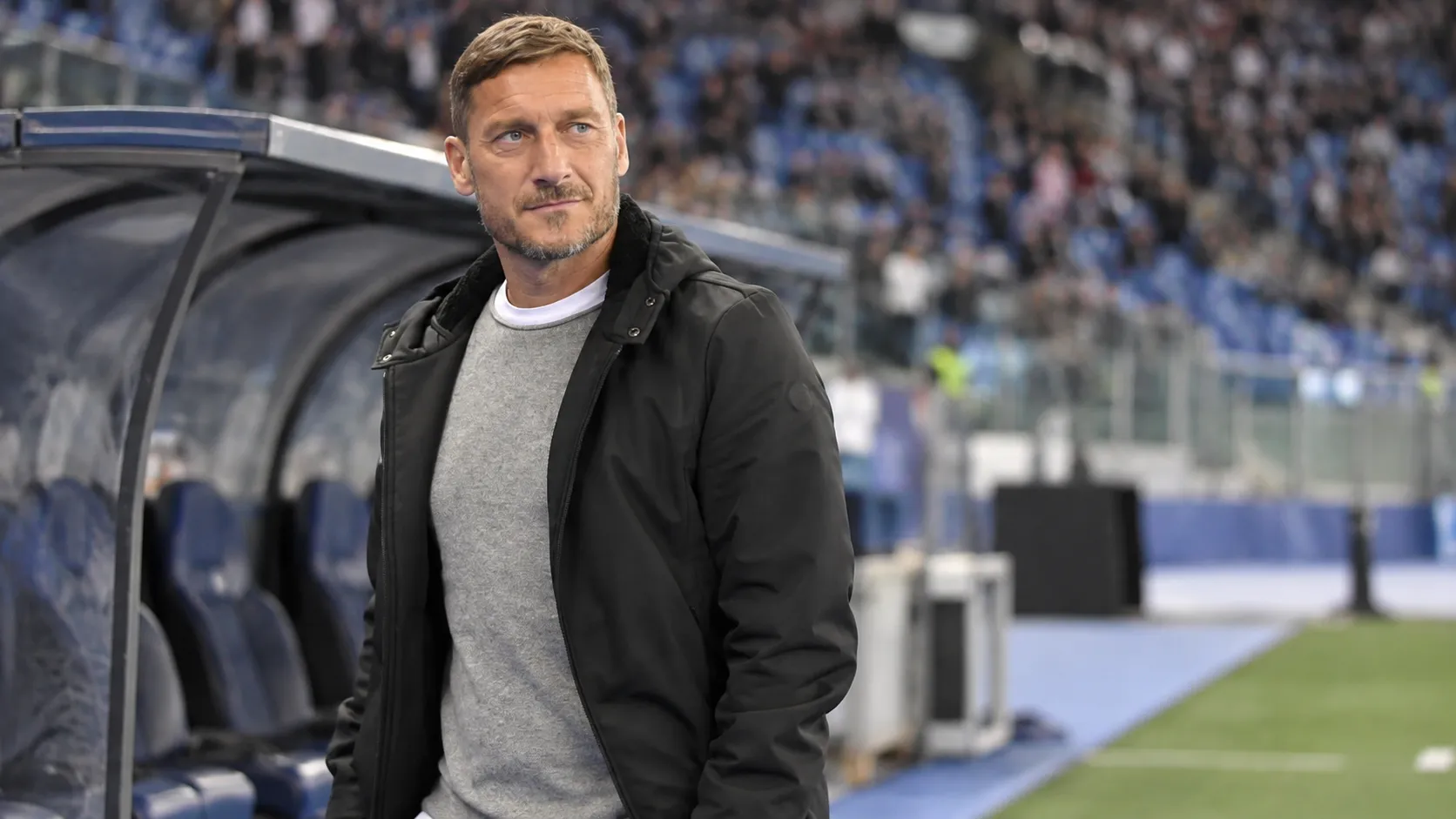 Francesco Totti staat rustig naast de dug-out in een voetbalstadion.