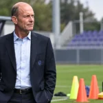 Een niet-spelende clubvertegenwoordiger in donker pak naast oefenmateriaal op het trainingsveld.