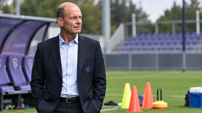 Een niet-spelende clubvertegenwoordiger in donker pak naast oefenmateriaal op het trainingsveld.