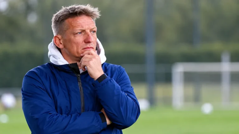 Wesley Sonck in een blauwe jas naast een voetbalveld.