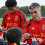Twee spelers in rode trainingskleding begroeten supporters aan een trainingsveld.