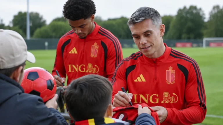 Twee spelers in rode trainingskleding begroeten supporters aan een trainingsveld.