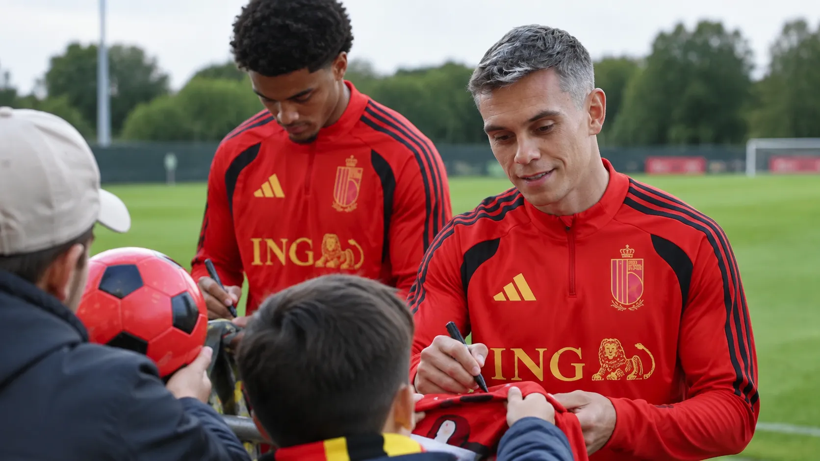 Twee spelers in rode trainingskleding begroeten supporters aan een trainingsveld.