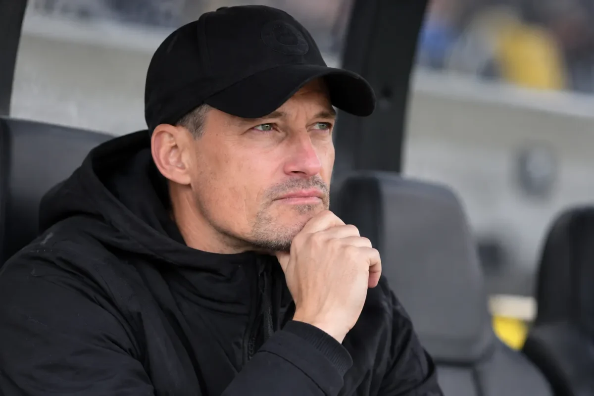 Alexander Blessin zit in zwarte coachingkleding op een reservebank in een stadion.