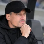 Alexander Blessin zit in zwarte coachingkleding op een reservebank in een stadion.