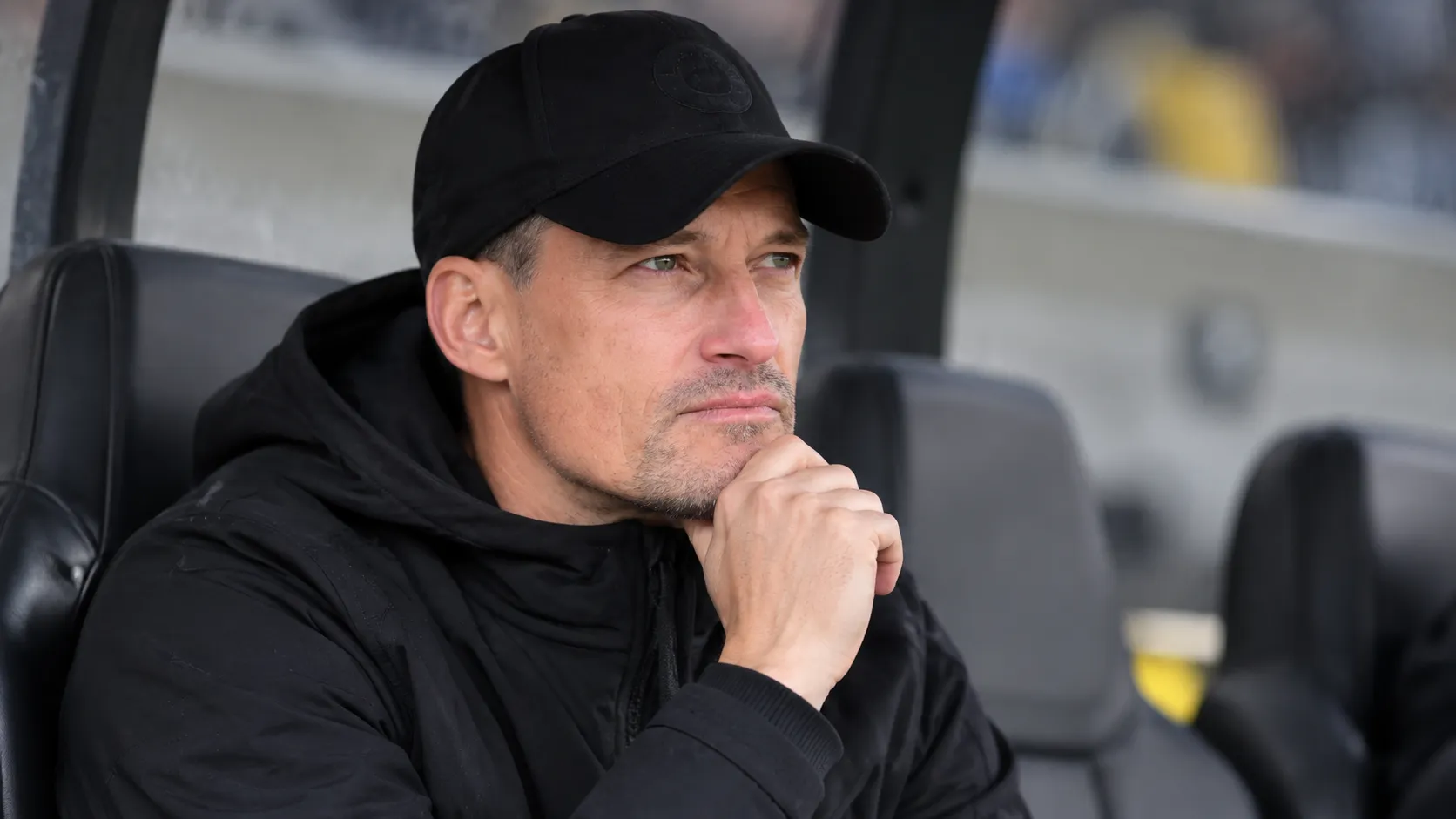 Alexander Blessin zit in zwarte coachingkleding op een reservebank in een stadion.