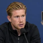 Kevin De Bruyne spreekt in een mediazone in een donkere trainingsjas voor een microfoon.