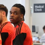 Lucas Stassin in rode en zwarte Belgische trainingskleding tussen ploegmaats in een indoorruimte.
