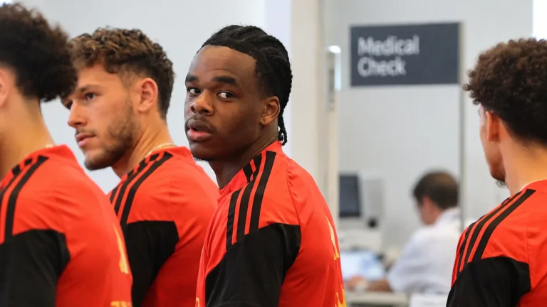 Lucas Stassin in rode en zwarte Belgische trainingskleding tussen ploegmaats in een indoorruimte.