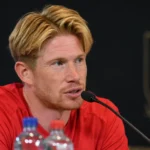 Kevin De Bruyne spreekt tijdens een persconferentie in een rood sportshirt.