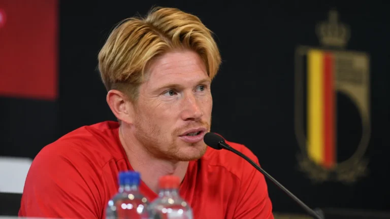 Kevin De Bruyne spreekt tijdens een persconferentie in een rood sportshirt.