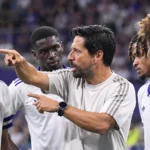 Een coach geeft aanwijzingen aan spelers langs de zijlijn van een voetbalstadion.
